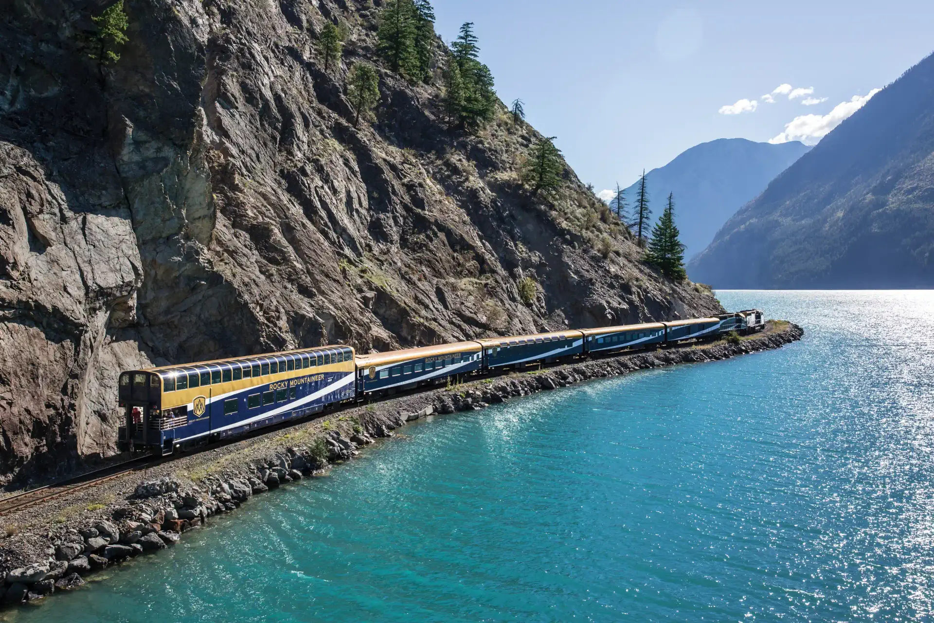 Scenic view of the Rocky Mountaineer train traveling along a rocky coastline beside a vibrant turquoise lake, surrounded by mountains and pine trees under a clear blue sky.